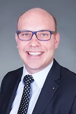 Ein Mann mit Brille in einem Anzug, lächelt für ein Porträt.