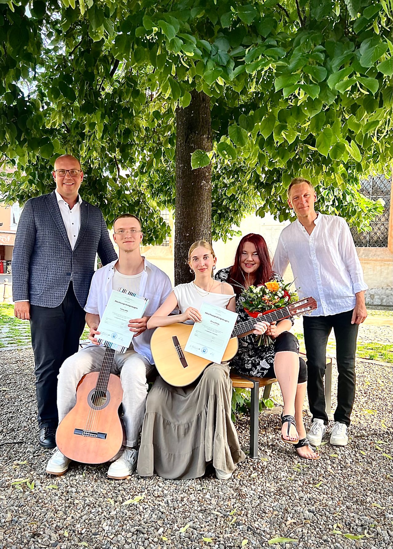 Fünf Menschen posieren lächelnd unter einem Baum für ein Foto. Drei halten Urkunden in der Hand und eine Frau hält eine Gitarre. Eine Frau sitzt auf einer Bank und zwei Männer stehen neben ihr.