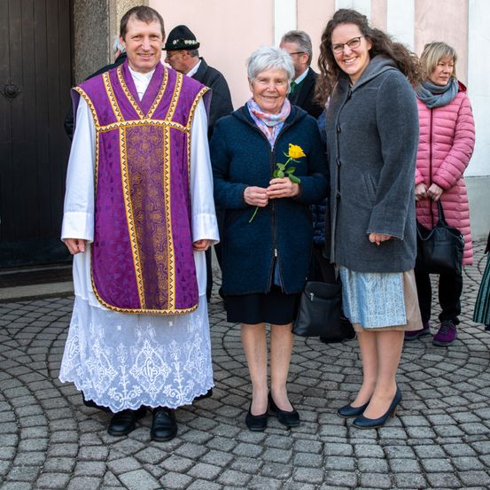 Ein Priester in liturgischen Gewändern steht mit zwei Frauen, eine hält eine gelbe Rose, vor einem Gebäude mit Zuschauern.