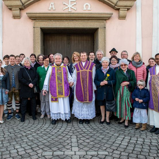 Eine Gruppe von Menschen, einschließlich Priester, steht vor einer Kirche und lächelt für ein Foto. Einige halten Blumen.