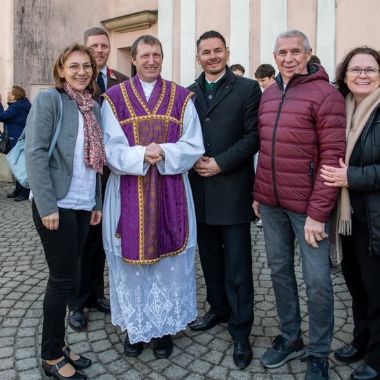 Ein Priester in traditioneller Kleidung steht mit drei Männern und zwei Frauen und lächelt für ein Foto. Dahinter sind mehrere Personen zu sehen, und ein Gebäude mit einer Ziegelmauer ist im Hintergrund.
