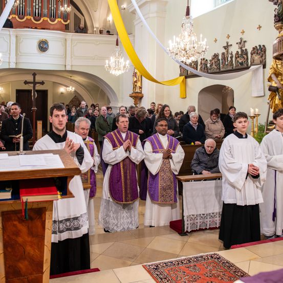Mehrere Priester stehen in einer Kirche und blicken zum Altar. Sie sind in traditionelle Roben gekleidet, wobei zwei von ihnen violette Stolen tragen. Hinter ihnen beobachtet eine Menge Menschen. Der Altar hat ein Kreuz und eine Kristallleuchter.