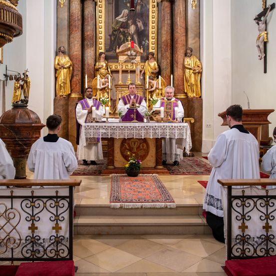 Eine Gruppe von Priestern in weißen Gewändern führt eine religiöse Zeremonie in einer Kirche durch. Sie stehen um einen Tisch mit Kerzen und Blumen, mit Statuen und einem Kruzifix im Hintergrund.
