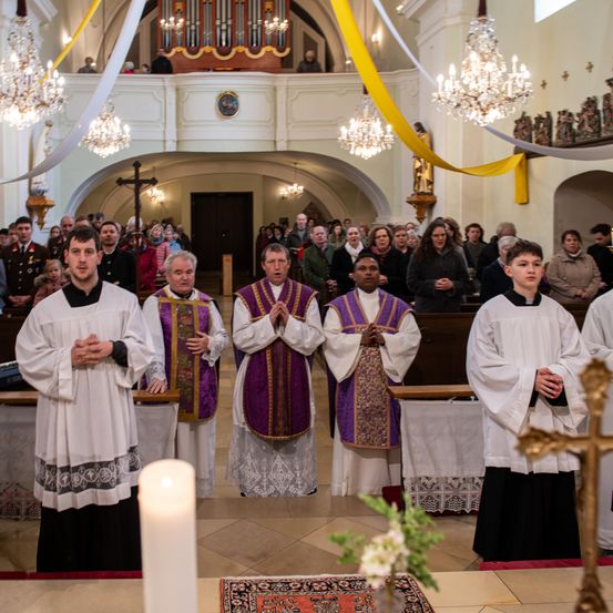 Priester stehen am Altar in einer Kirche, mit Kerzen und Blumen. Eine Gemeinde sitzt hinter ihnen, und ein Militäroffizier ist anwesend.