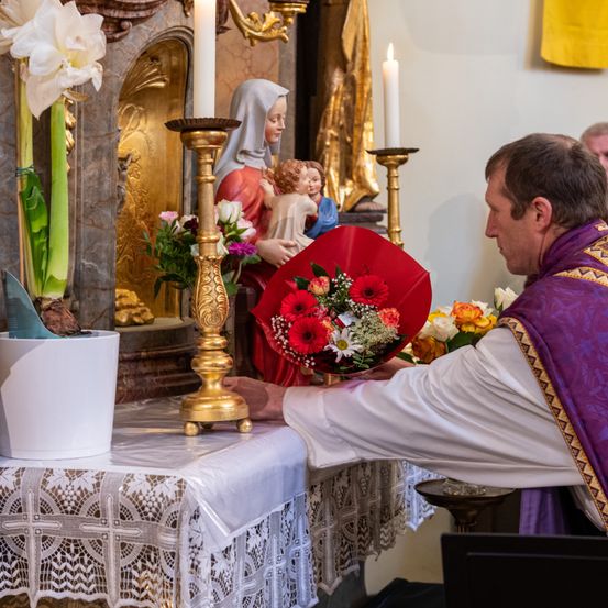 Ein Priester legt einen Blumenstrauß auf einen Altar in einer Kirche, mit einer Statue von Maria und Jesus hinter ihm.
