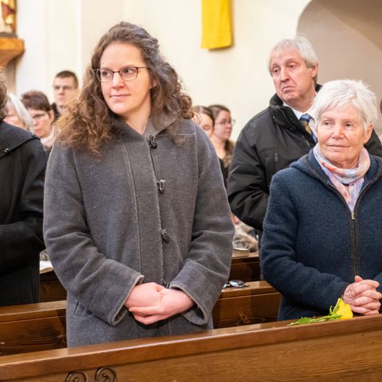 Eine Gruppe von Menschen versammelt sich in einer Kirche. Eine Frau mit Brille und lockigem Haar steht in der Mitte und sieht geradeaus. Eine ältere Frau mit weißen Haaren steht neben ihr und hält eine gelbe Blume. Dahinter sitzen andere Menschen auf Holzbänken.