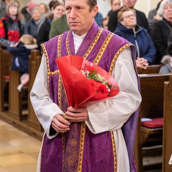 Ein Priester steht in einer Kirche und hält einen Strauß roter Blumen, während Menschen auf den Kirchenbänken hinter ihm sitzen.