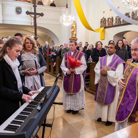 Eine Pianistin spielt bei einem Gottesdienst, umgeben von Gemeindemitgliedern und Priestern, einer hält Blumen.