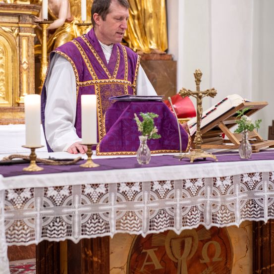 Ein Priester sitzt am Altar, mit einem violetten Tuch und zwei brennenden Kerzen. Vor ihm stehen ein Kreuz, Bücher und eine kleine Pflanze. Hinter ihm ist eine goldene Statue, und die Wand ist weiß.