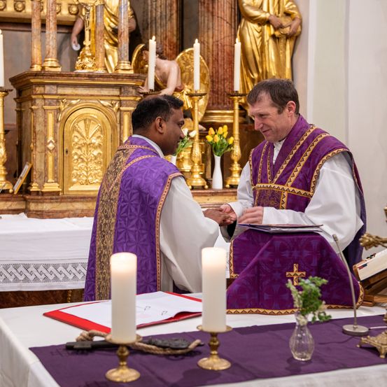 Zwei Priester in violetten Gewändern tauschen einen Händedruck in einer Kirche aus. Dahinter befindet sich ein Altar mit Kerzen und Statuen. Ein Buch und Blumen liegen auf dem Tisch.