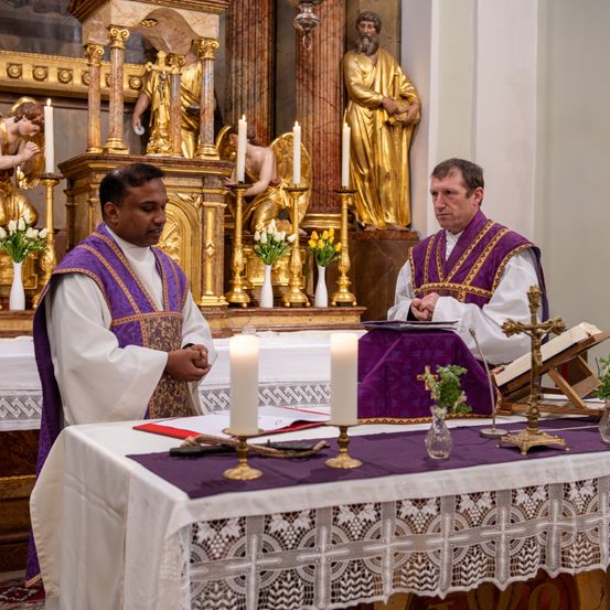 Zwei Priester in lila Gewändern stehen vor einem Altar in einer Kirche. Sie beten mit Kerzen und Büchern auf dem Altar vor sich. Hinter ihnen stehen Statuen von Engeln und einem Heiligen auf dem Altar.