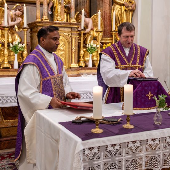 Zwei Priester stehen in einer Kirche an einem Tisch, einer liest aus einem Buch und der andere sieht sich ein Dokument an. Hinter ihnen schmücken goldene Statuen und Kerzen den Altar.
