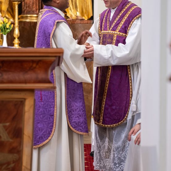 Zwei Priester schütteln sich die Hände in einer Kirche. Einer trägt ein weißes Gewand und der andere ein lilafarbenes. Hinter ihnen steht ein goldener Kerzenständer.