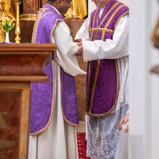 Zwei Priester in lila und weißen Gewändern schütteln sich die Hände in einer Kirche. Hinter ihnen steht eine Statue und eine Blumenvase.