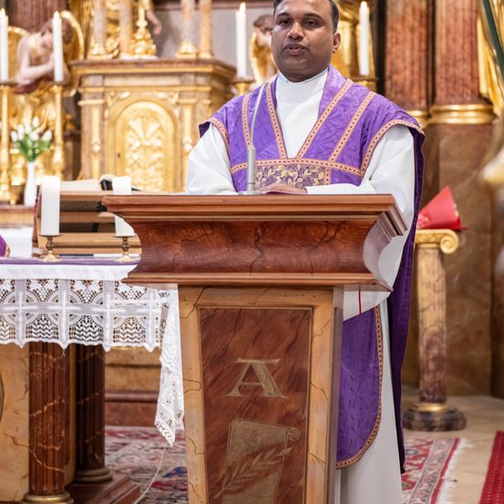 Ein Priester steht in einer Kirche an einer Kanzel, gekleidet in violette und weiße Gewänder, mit Kerzen und einer Blumendekoration im Hintergrund.