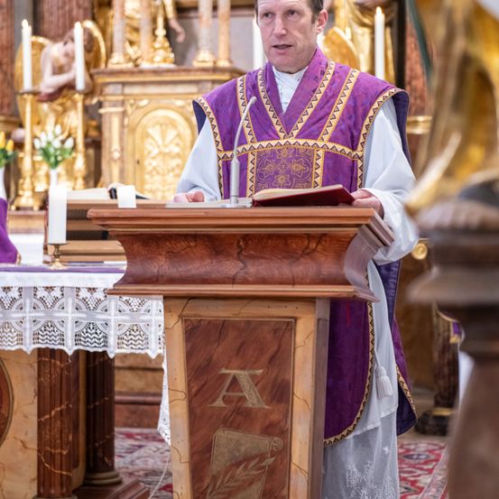 Ein Priester steht an einer hölzernen Kanzel in einer Kirche, liest aus einem Buch, mit kunstvollen Dekorationen und Kerzen im Hintergrund.