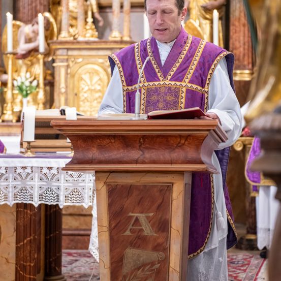 Ein Priester steht an einem Pult und liest aus einem Buch mit einem Mikrofon. Er trägt ein violettes und weißes Gewand. Hinter ihm steht ein goldener Altar mit Statuen und Kerzen.