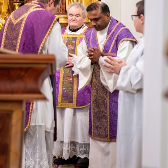 Priester in traditioneller Kleidung stehen in einer Kirche, einer hält die Hände zum Gebet gefaltet. Links ist ein hölzerner Predigtstuhl zu sehen.