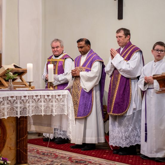 Vier Priester stehen in einer Kirche und beten mit gefalteten Händen. Vor ihnen steht ein Tisch mit weißer Spitzendecke und Kerzen. Ein Junge mit Brille steht rechts und ein Kreuz ist an der Wand befestigt.