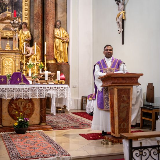 Ein Priester steht in einer Kirche an einer Kanzel, mit goldenen Statuen und einem Kruzifix an der Wand hinter ihm. Vor ihm steht ein Altar, geschmückt mit Blumen und Kerzen.