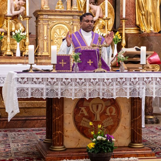 Ein Priester steht am Altar, trägt eine purpurne Robe und erhebt die Hände. Hinter ihm stehen Statuen und goldene Kerzen. Der Altar ist mit Blumen und einem Kreuz geschmückt.