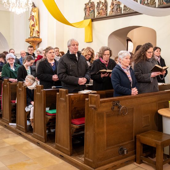 Eine Gruppe von Menschen unterschiedlichen Alters versammelt sich in einer Kirche und betet mit gefalteten Händen. Einige stehen, andere sitzen in hölzernen Kirchenbänken. Die Kirche ist mit dekorativen Elementen wie Statuen und einem Bogen ausgestattet.