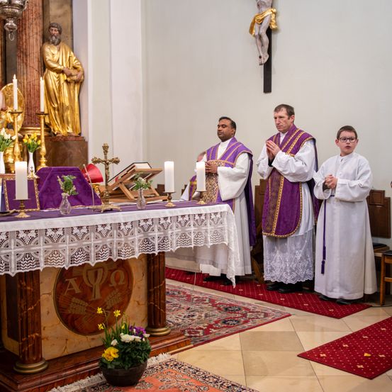 Drei Priester stehen an einem Altar in einer Kirche, mit einem Jungen davor. Eine goldene Statue eines Heiligen steht hinter ihnen, und der Altar ist mit Kerzen, Blumen und einem Kruzifix geschmückt.