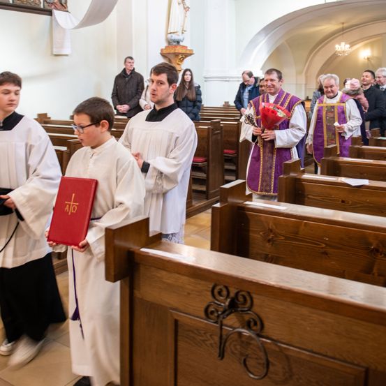 In einer Kirche versammeln sich mehrere Personen für eine Zeremonie. Ein Junge hält ein rotes Buch, und zwei Männer in weißen Gewändern stehen vor ihm. Ein älterer Mann hält einen Blumenstrauß.