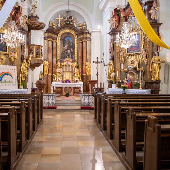 Ein prächtiger Kircheninnenraum mit einem prächtigen Altar, goldenen Statuen, einer zentralen Kristallleuchter und einem bunten Banner. Die Kirchenbänke sind in Reihen angeordnet und führen zum Altar. Der Altar ist von Kerzen umgeben und hat eine Blumenarrangement.