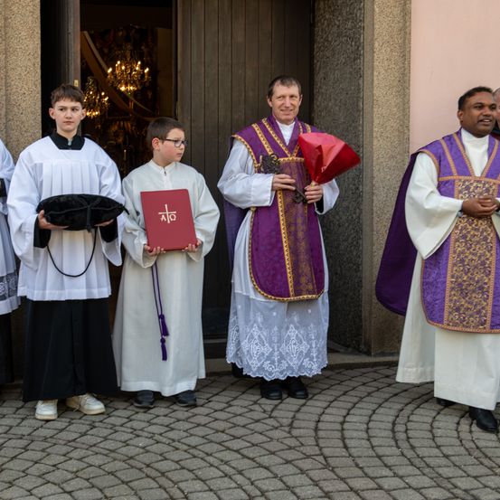 Mehrere Geistliche stehen vor einer Kirche. Ein Priester hält einen roten Blumenstrauß. Ein Junge hält ein rotes Buch mit einem Kreuzsymbol.