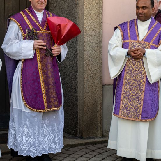 Zwei Priester in aufwendigen Gewändern stehen im Freien, einer hält einen roten Blumenstrauß und der andere hat die Hände gefaltet. Sie stehen in der Nähe einer Steinmauer.