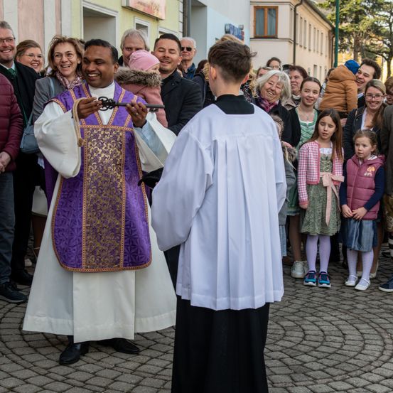 Ein Priester in einer violetten Tunika hält einen Schlüssel bei einer Zeremonie, während ein junger Junge in einer weißen Robe zusieht, umgeben von einer Menschenmenge auf einer Kopfsteinpflasterstraße.