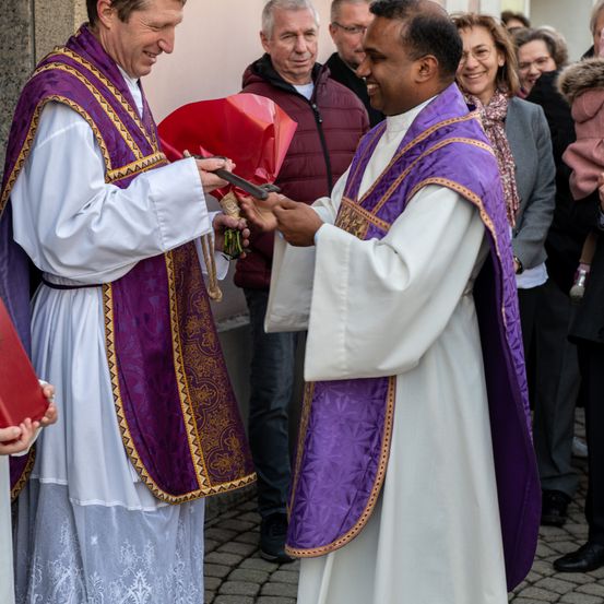 Zwei Priester in weißen und lila Gewändern tauschen draußen Blumen aus. Hinter ihnen stehen einige Leute und beobachten die Veranstaltung.