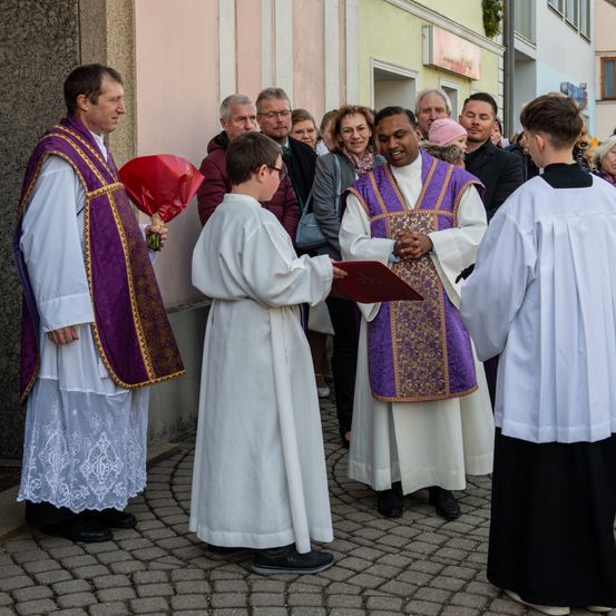 Priester in zeremonieller Kleidung stehen vor einem Gebäude, ein junger Junge hält ein Buch und einen Blumenstrauß. Dahinter beobachten mehrere Personen.