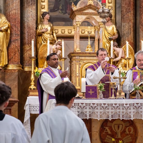 Zwei Priester führen eine religiöse Zeremonie in einer Kirche durch. Sie halten Kerzen und ein Buch. Eine goldene Statue eines Engels steht hinter ihnen.