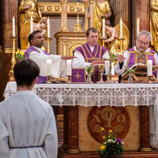 Drei Priester führen eine Zeremonie an einem Altar durch. Einer liest aus einem Buch, ein anderer gestikuliert und der dritte hält einen Kelch. Ein Gläubiger steht im Vordergrund. Der Altar ist mit Kerzen und Blumen geschmückt. Hinter den Priestern befinden sich goldene Engelsstatuen.