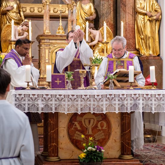 Drei Priester führen eine Zeremonie an einem Kirchenaltar durch, umgeben von goldenen Statuen und Kerzen. Ein Priester hält ein weißes Tuch, während ein anderer aus einem Buch liest. Ein Anbeter beobachtet von der Seite.