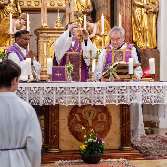 Drei Priester stehen an einem Altar und führen eine Zeremonie mit Kerzen und einem Kelch in einer Kirche durch. Eine Person kniet davor und Blumen schmücken den Altar.