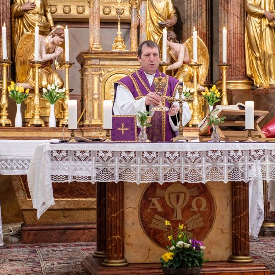 Ein Priester steht vor einem Altar, hält ein goldenes Kreuz und steht vor einem Tisch mit Kerzen und Blumen. Dahinter befinden sich Statuen von Engeln und ein goldener Hintergrund.