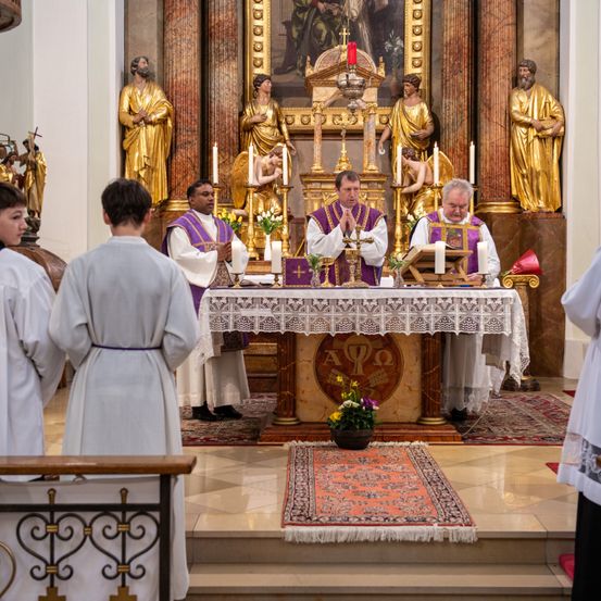 Zwei Priester führen eine Zeremonie an einem Altar mit drei Altarjungen durch, während andere zusehen. Dahinter befinden sich Statuen von Männern und ein Gemälde.