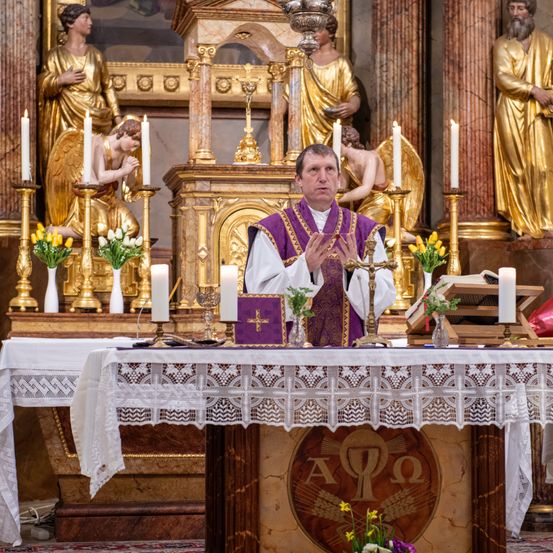 Ein Priester steht in einer Kirche am Altar, umgeben von Kerzen und Blumen. Im Hintergrund befinden sich goldene Statuen von Engeln und Cherubinen.