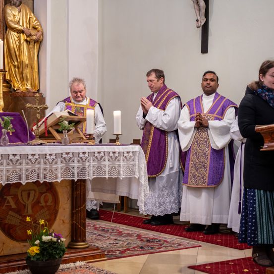 In einer Kirche stehen vier Priester vor einem Altar. Der Altar hat ein Kreuz, Kerzen und ein Buch. Dahinter steht eine Statue eines Heiligen.
