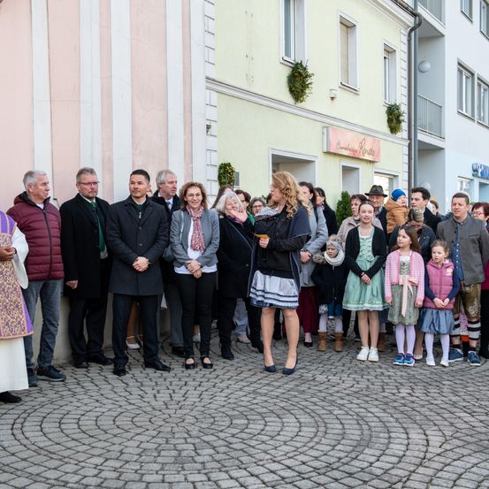 Eine Gruppe von Menschen steht auf einer gepflasterten Straße, einige in formeller Kleidung und andere in lässiger Kleidung, mit zwei jungen Mädchen in Kleidern vorne. Dahinter ist ein Gebäude mit einem Schild und einem festlichen Kranz zu sehen.