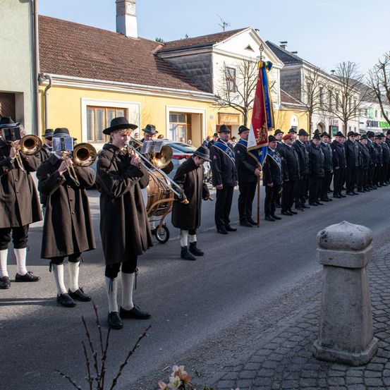 Eine Marschkapelle steht in Formation auf einer Straße, wobei die Musiker Trompeten und Posaunen spielen. Die Band ist in traditioneller Kleidung gekleidet. Im Hintergrund sind Gebäude mit braunen Dächern zu sehen.