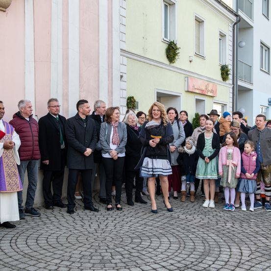 Eine Gruppe von Menschen steht vor einem Gebäude mit rosa Wänden und gelben Fenstern. Eine Frau in der Mitte lächelt und hält ein Buch. Einige tragen Winterkleidung, andere Kleider. Ein Priester steht links.