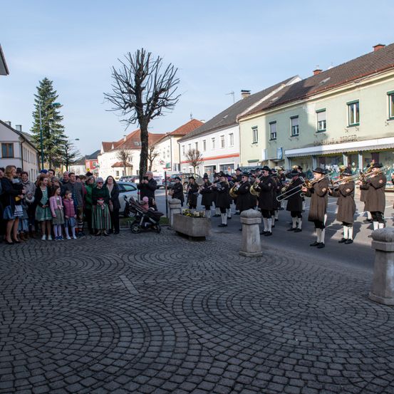Ein Blasorchester spielt auf einer Kopfsteinpflasterstraße, während eine Menschenmenge zusieht. Im Hintergrund befinden sich Gebäude und Bäume.