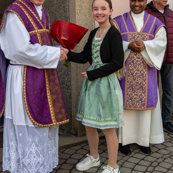 Drei Priester in traditioneller Kleidung stehen mit einem jungen Mädchen in einem grünen Kleid, das einen roten Blumenstrauß hält, lächelnd auf einer Kopfsteinpflasterstraße.