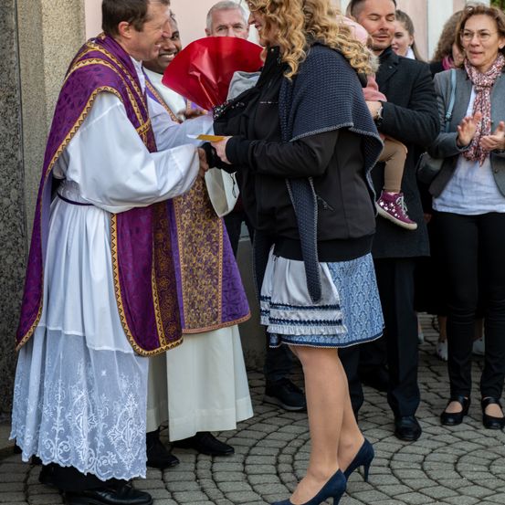 Zwei Priester reichen einer Frau in einer Kirche ein Dokument, wobei mehrere Menschen um sie herum zusehen, darunter eine Frau mit Brille.