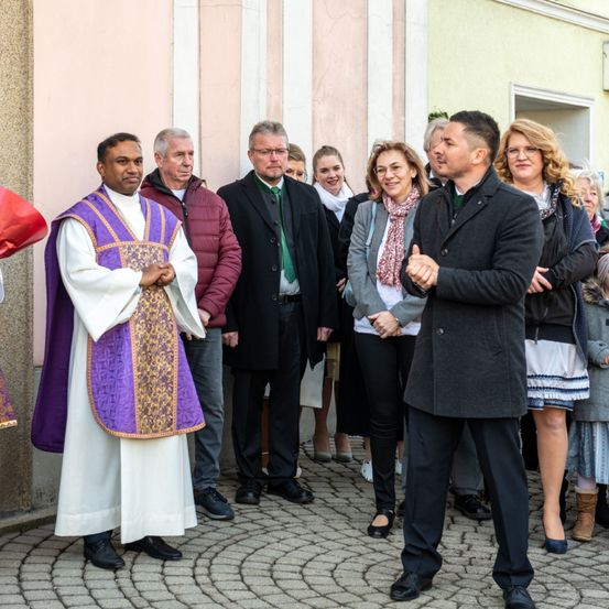 Ein Priester in einer weißen und lila Robe steht vor einer Gruppe von Menschen, die in Winterkleidung gekleidet sind. Hinter ihm deutet ein Mann in einem schwarzen Mantel, während er spricht.