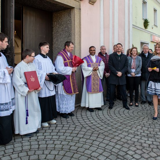 Eine Gruppe von Geistlichen und Gemeindemitgliedern steht vor einer Kirche. Sie tragen weiße und lila Roben und halten Bücher.